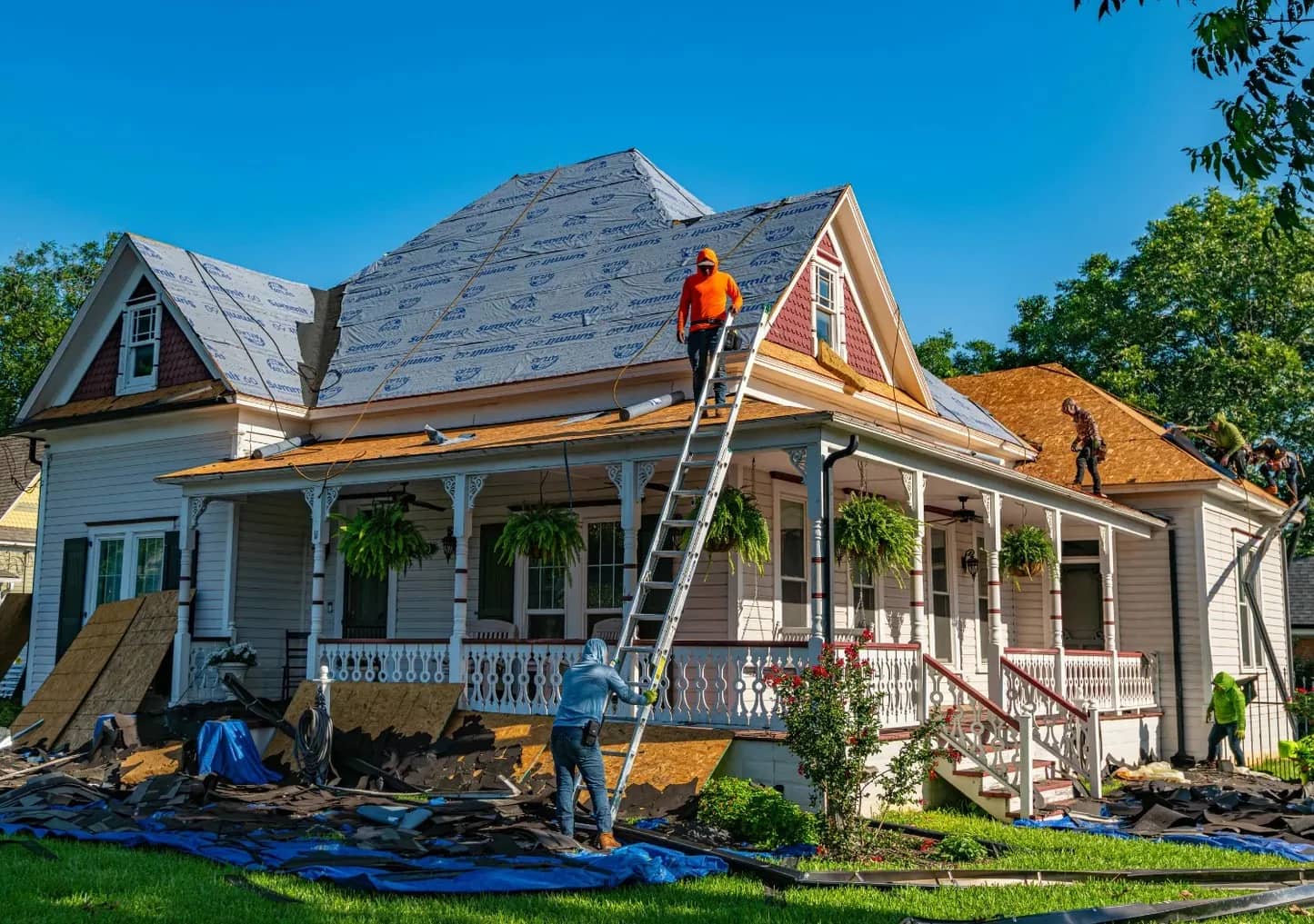 A RainCity crew re-roofing a heritage house on a clear day: one worker on a ladder at the ridge, others laying underlay across the porch roof, stripped shingles bagged on tarps below.