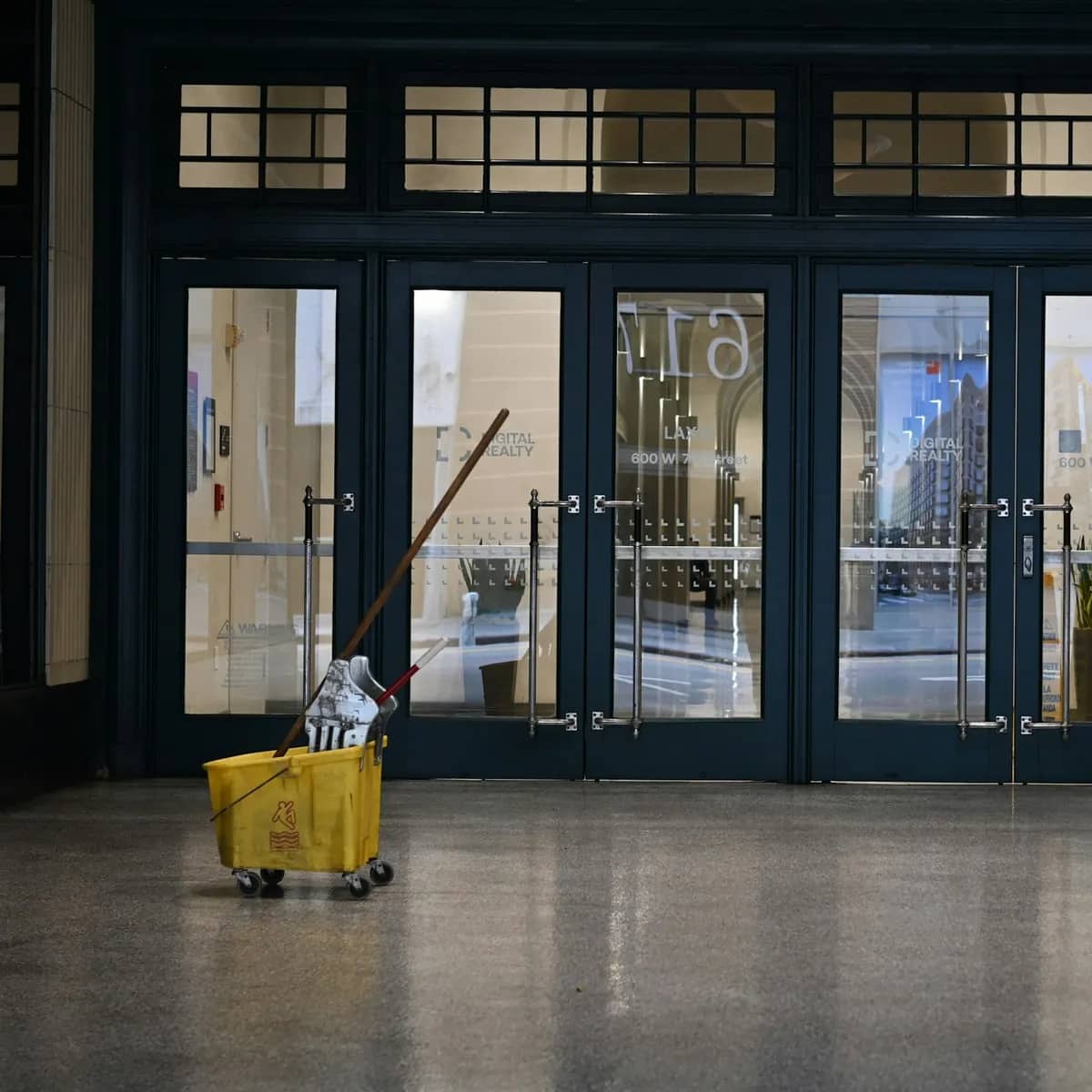 A yellow mop bucket inside the glass entrance doors of an office lobby, the polished stone floor still wet.