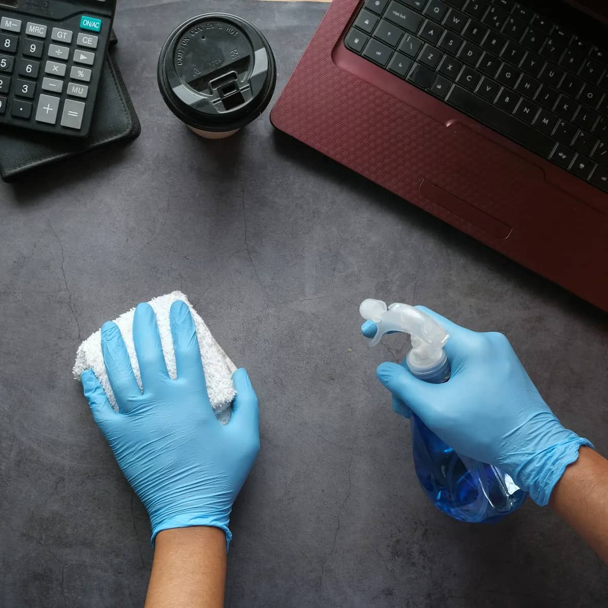 Gloved hands wiping a dark office desk with a cloth and a spray bottle, beside a laptop and a calculator.