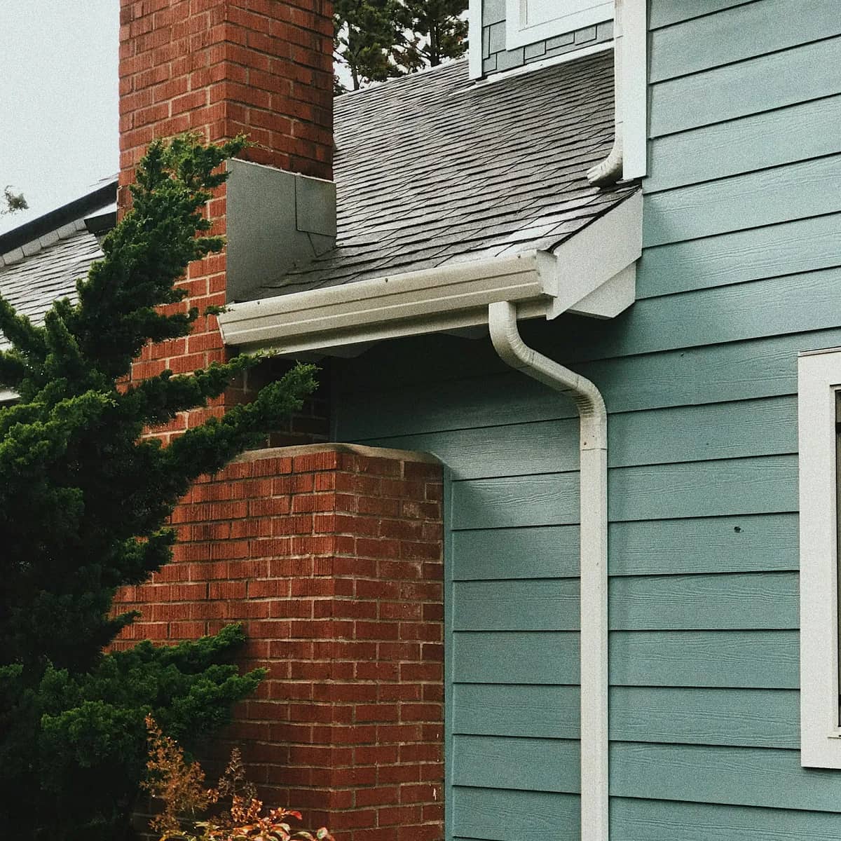 A white gutter and downspout running down pale blue siding beside a brick chimney.