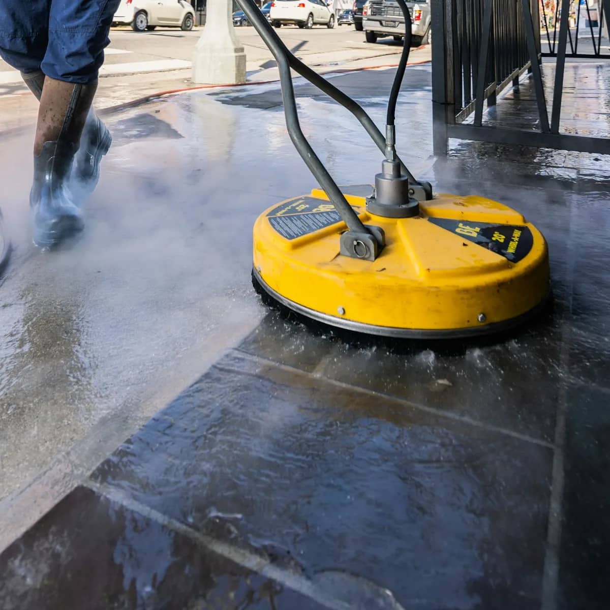 A rotary surface cleaner working across wet stone paving, the worker's boots and hose behind it.