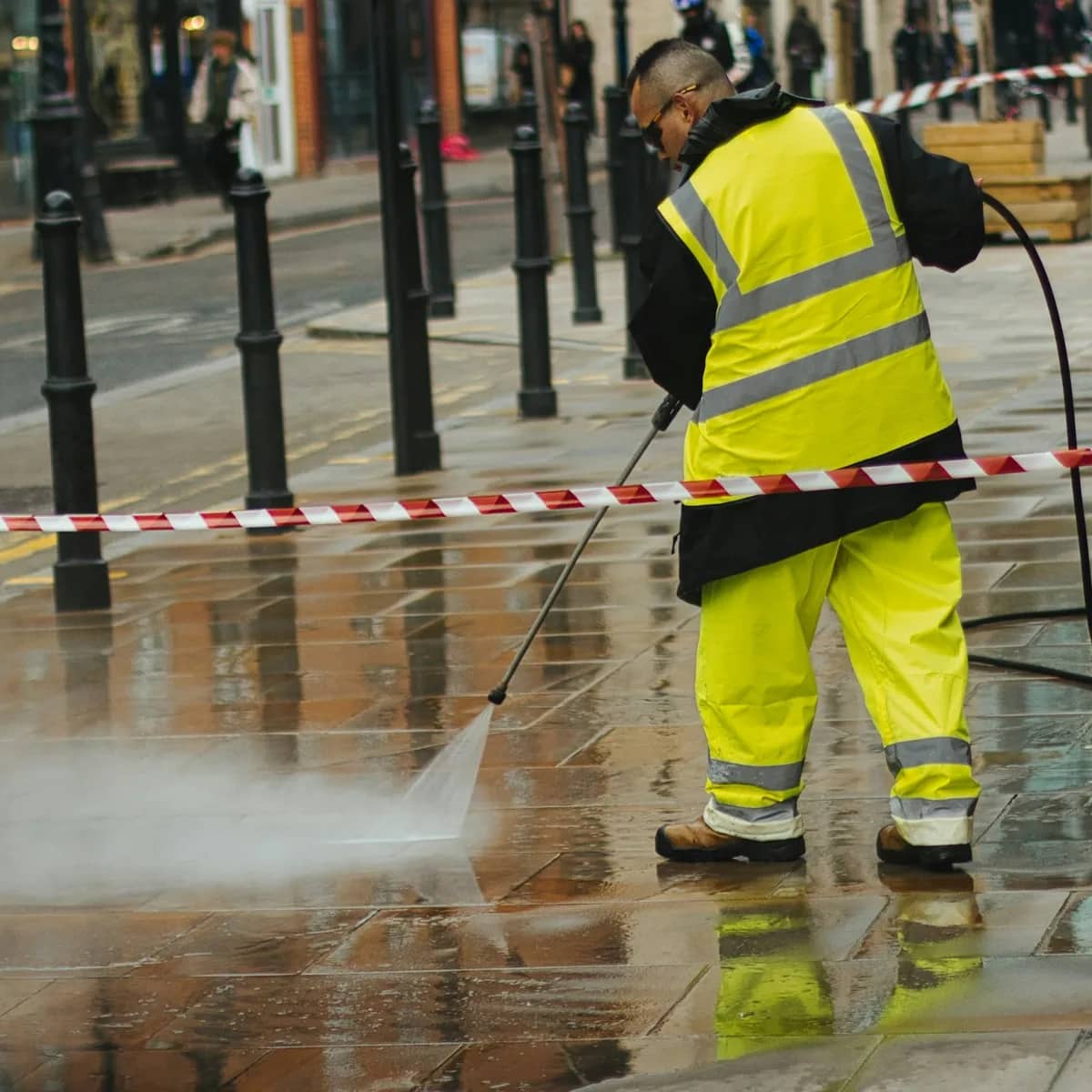 A worker in hi-vis pressure-washing a paved town sidewalk behind barrier tape, spray lifting off the stone.