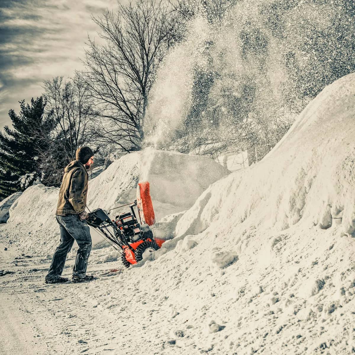 A man pushing an orange snow blower between high snowbanks, a plume of snow thrown into the air.