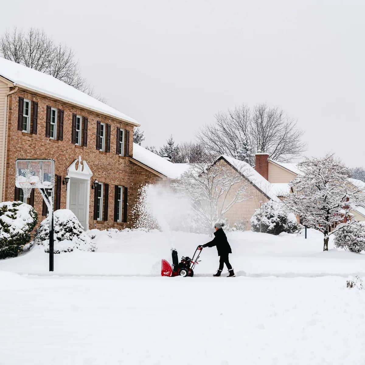 A person running a red snow blower along a snowy front walk in front of a brick house.