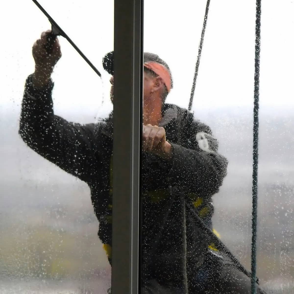 A window cleaner in a harness drawing a squeegee across wet glass, seen from inside through the pane.