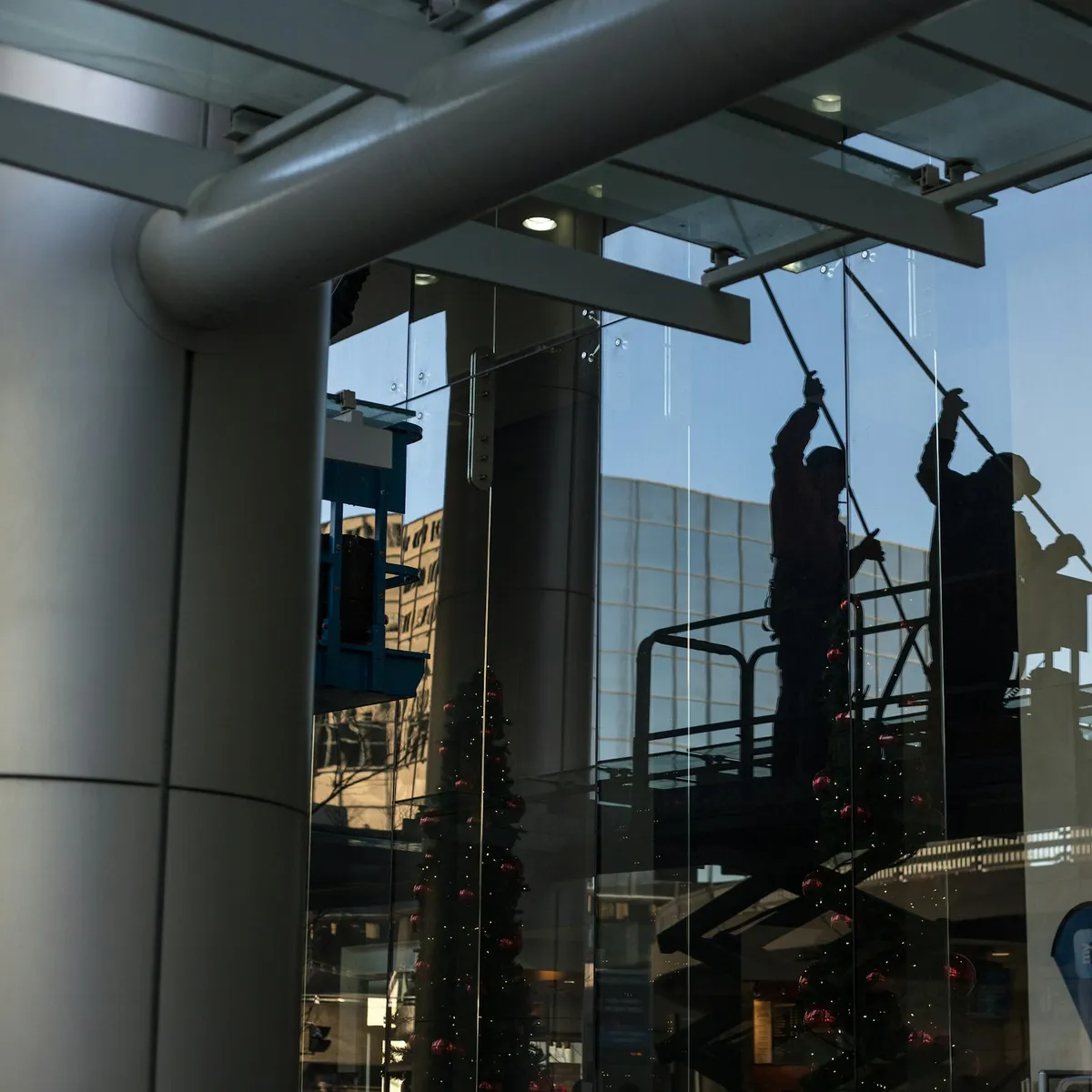 Two window cleaners on a lift working poles across a tall glass frontage, seen in silhouette.