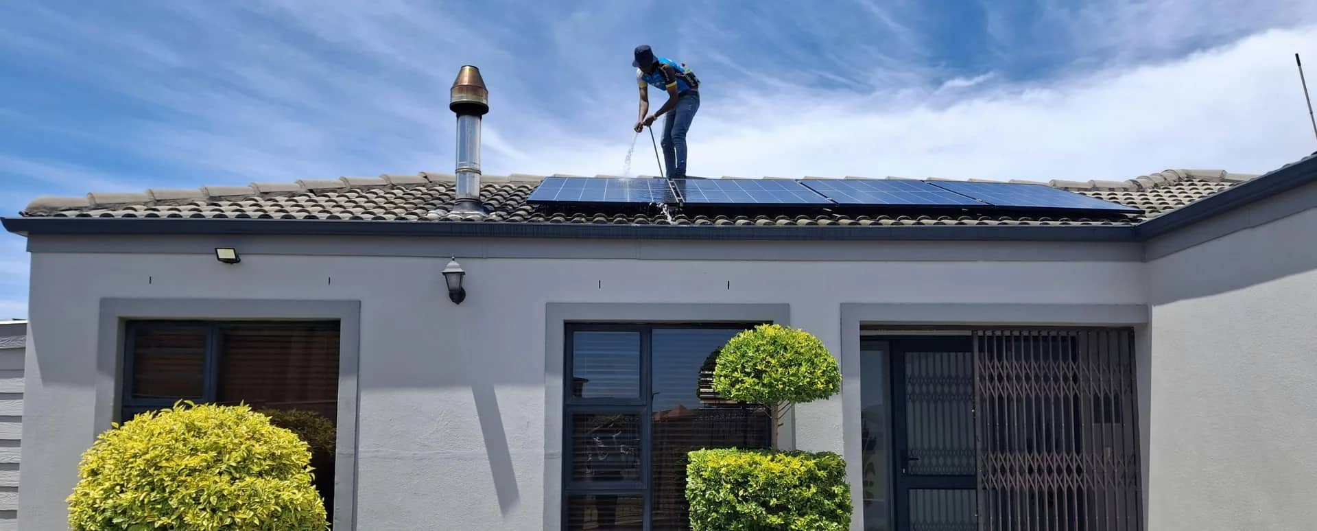 A RainCity technician on a tiled Greater Vancouver roof running a pressure-washing wand across a row of solar panels under a clear blue sky.