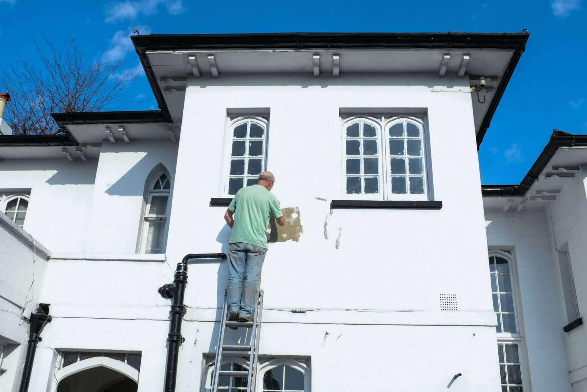 Painting: a decorator on a stepladder cutting in around a window on the white rendered exterior of a house.