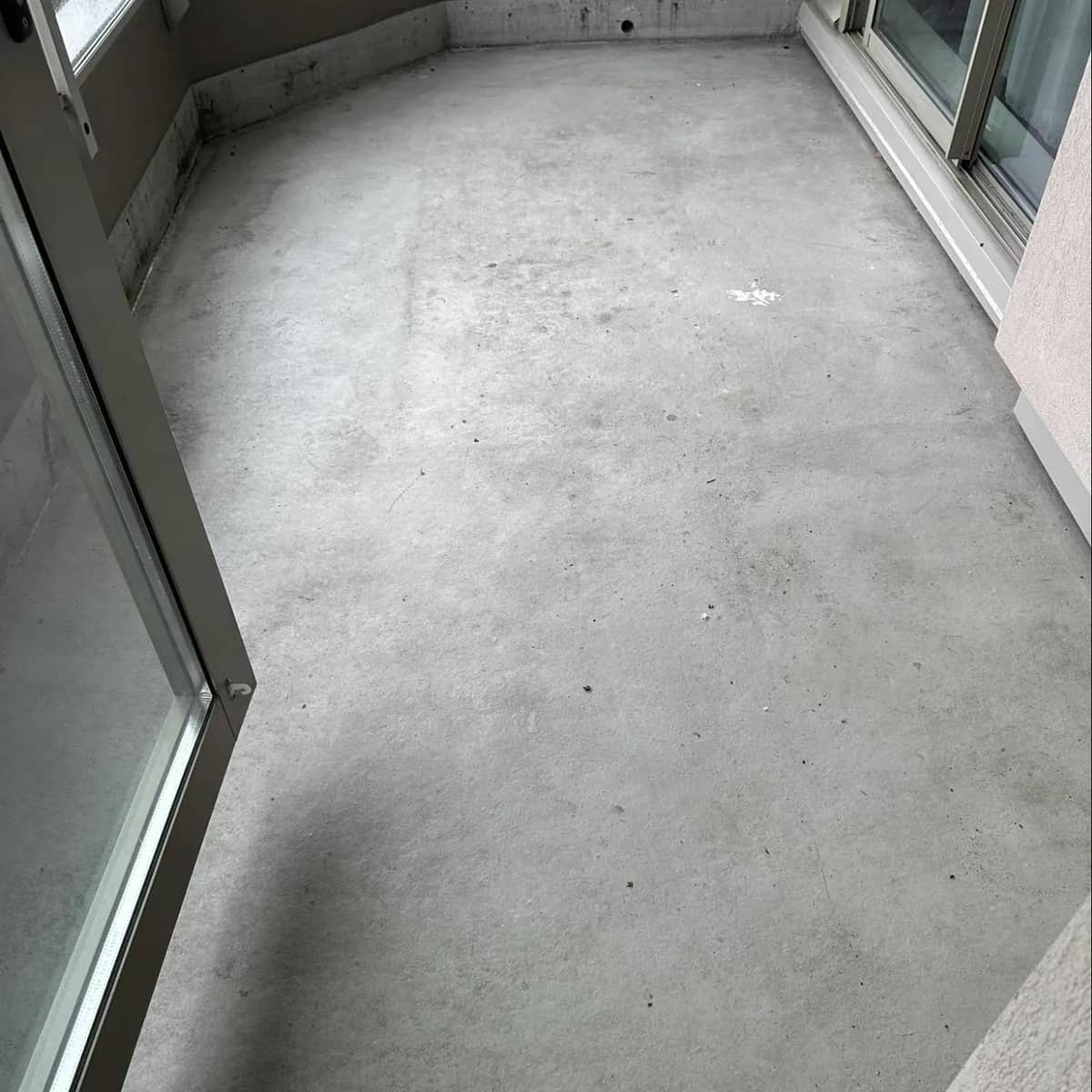An enclosed glass balcony before cleaning, its grey floor dull and stained, with a dirty tidemark along the base of the walls.