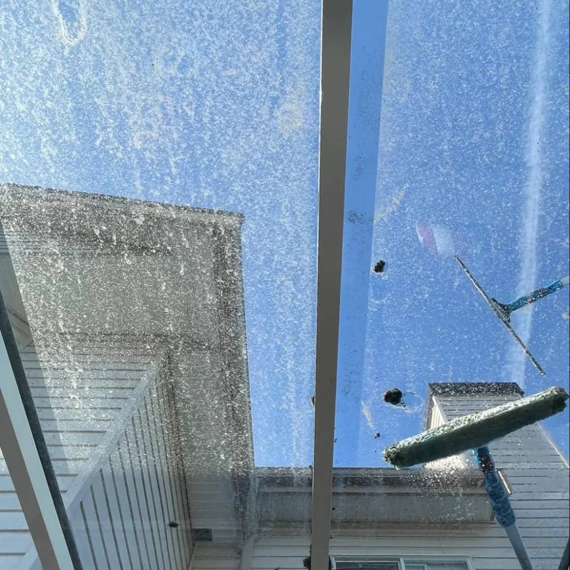 Looking up through a glass patio cover before cleaning: the panels spotted and filmed with grime, a squeegee on a pole working across them.