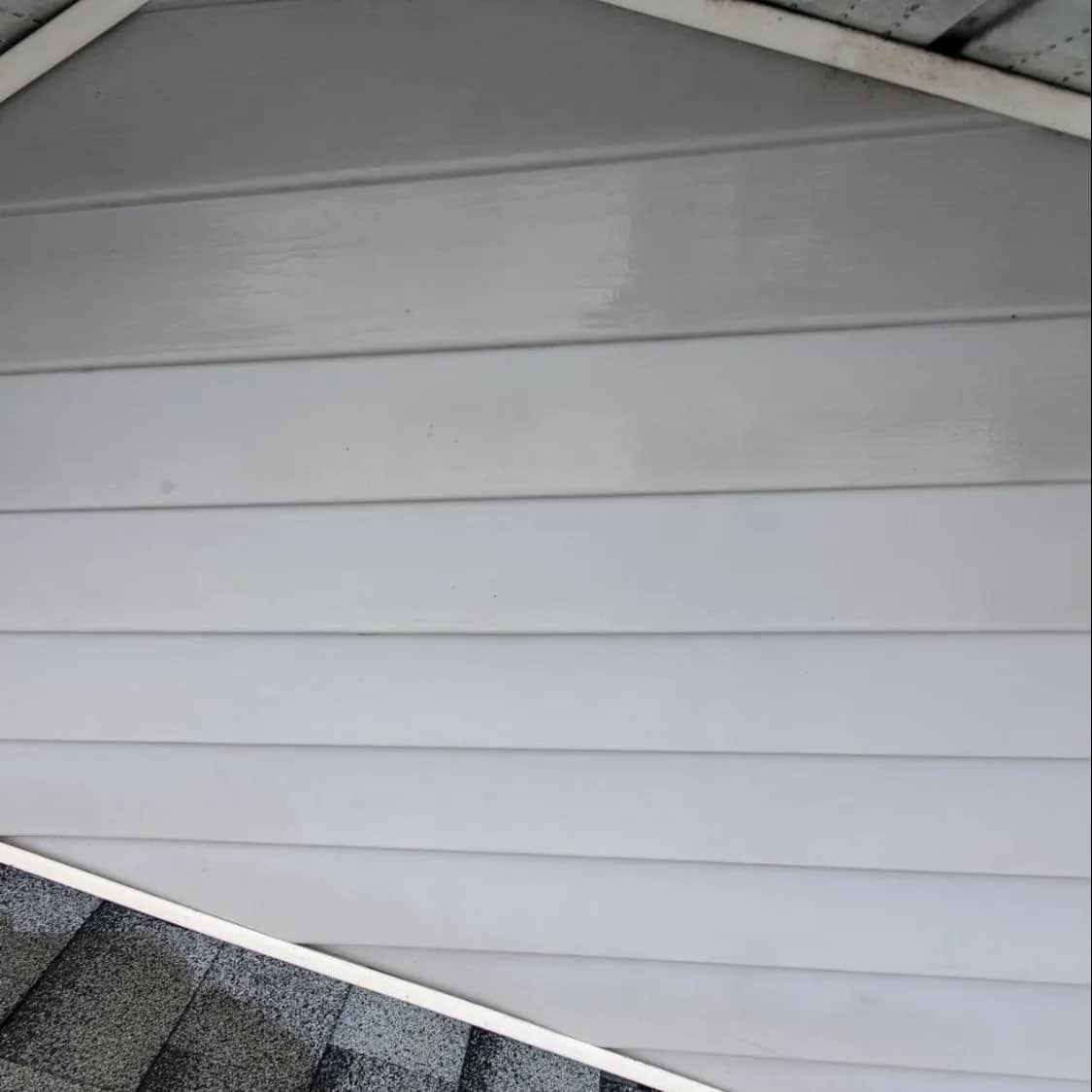 Horizontal siding and trim under a gable after washing, the boards clean and bright above a shingle roof.