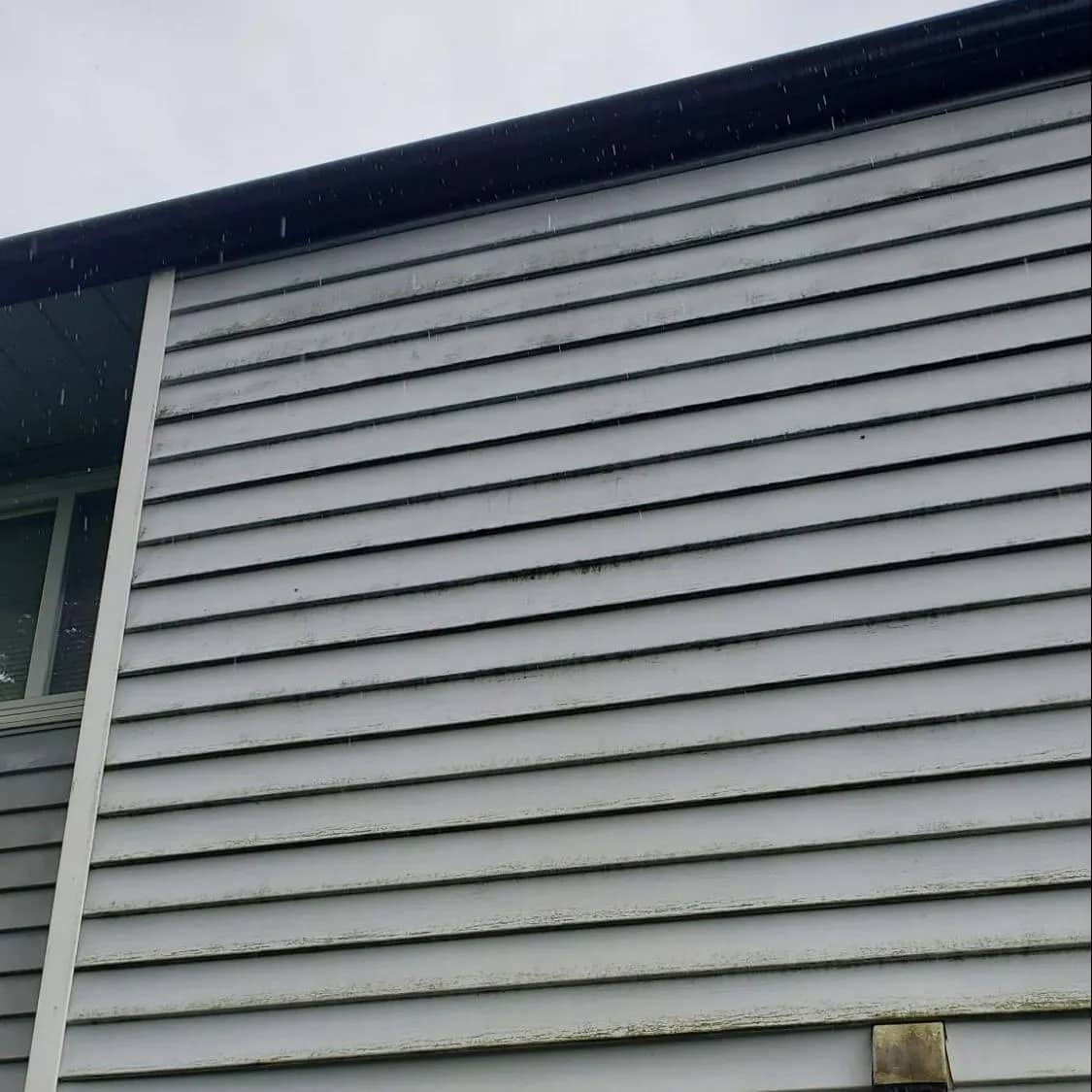 Pale grey horizontal siding under an eave before washing, streaked with dark algae and dirt along every board.