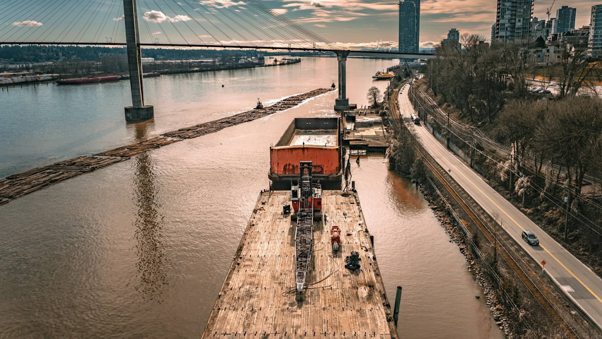 Downtown New Westminster from above the Fraser River: a work barge moored under the SkyBridge, log booms on the water, and the city's towers rising behind the rail line and Front Street.