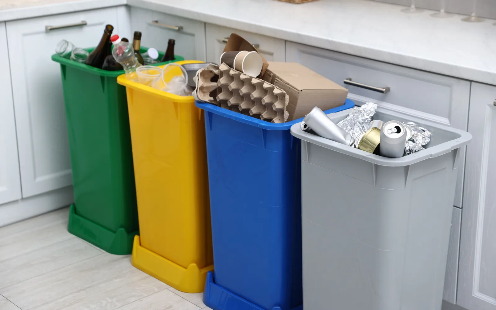 Commercial cleaning: four colour-separated recycling bins lined up against pale cabinetry, holding glass, plastics, flattened card and cans.