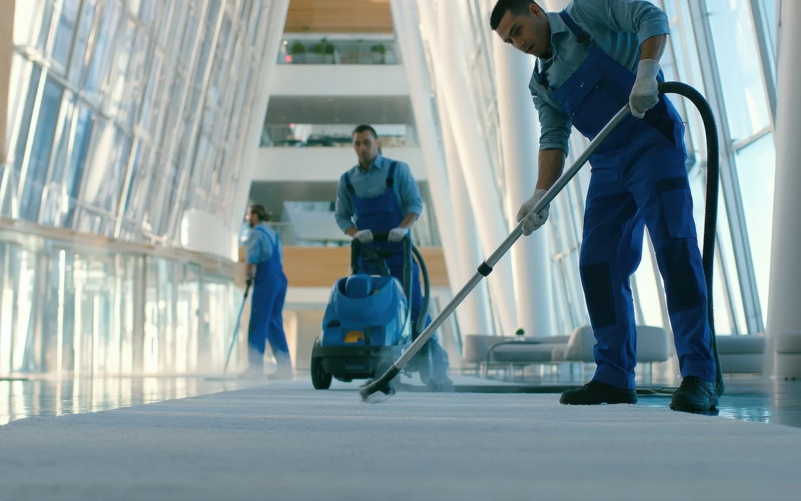 Commercial cleaning: three cleaners in blue uniforms working a carpeted atrium walkway, the nearest drawing a vacuum wand along the floor with a carpet machine running behind him.