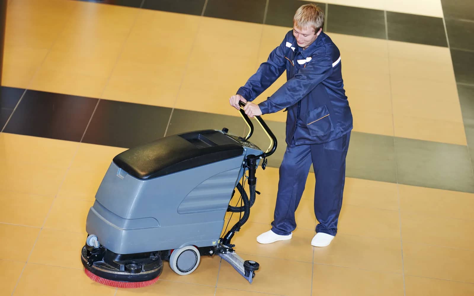 Commercial cleaning: an operative in navy overalls walking a scrubber-drier across a tiled floor, the machine's red brush head visible at the front of the deck.