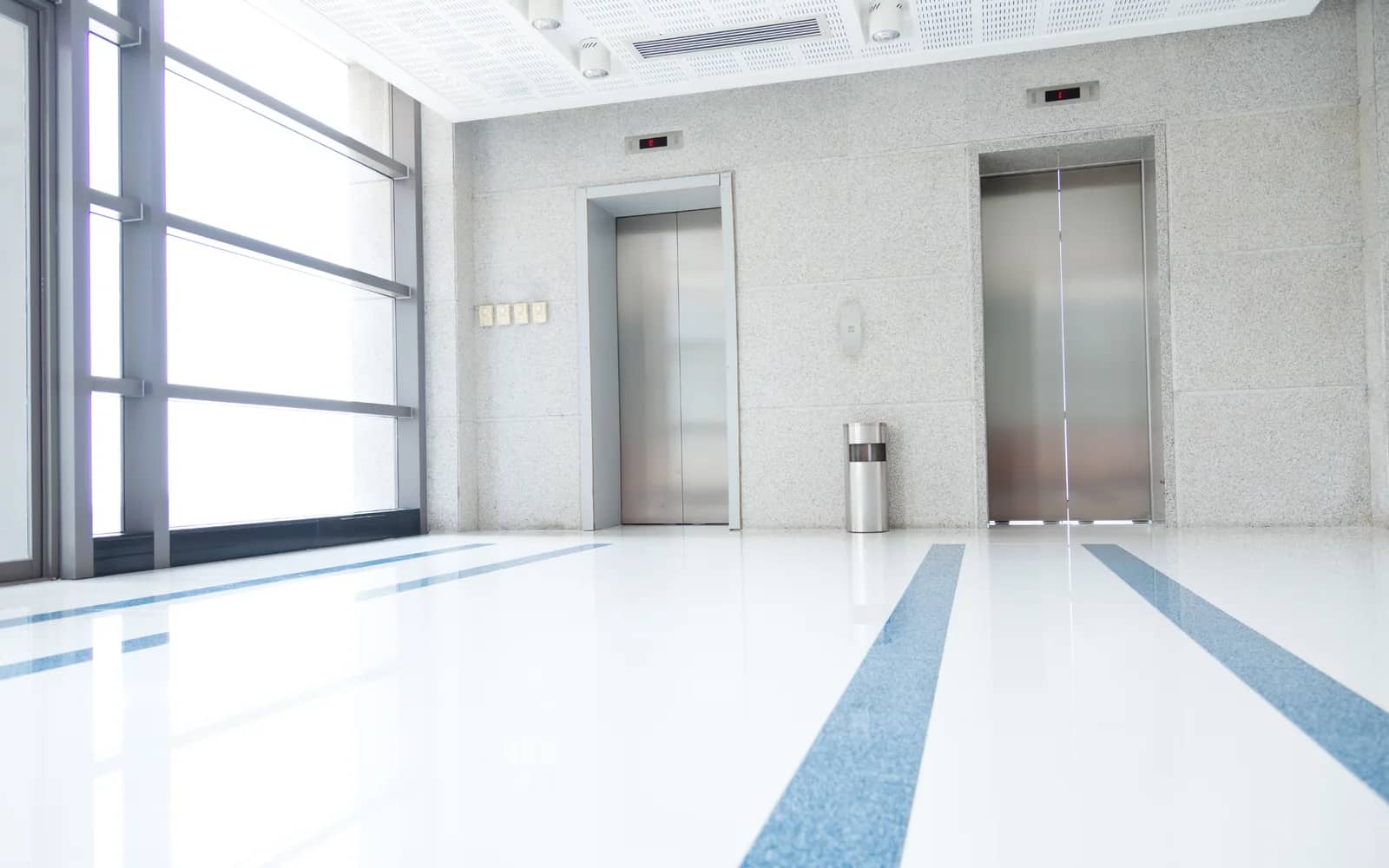 Commercial cleaning: an empty lift lobby with two stainless steel lift doors, the polished floor and its blue inlay stripes running unbroken to full-height glazing on the left.