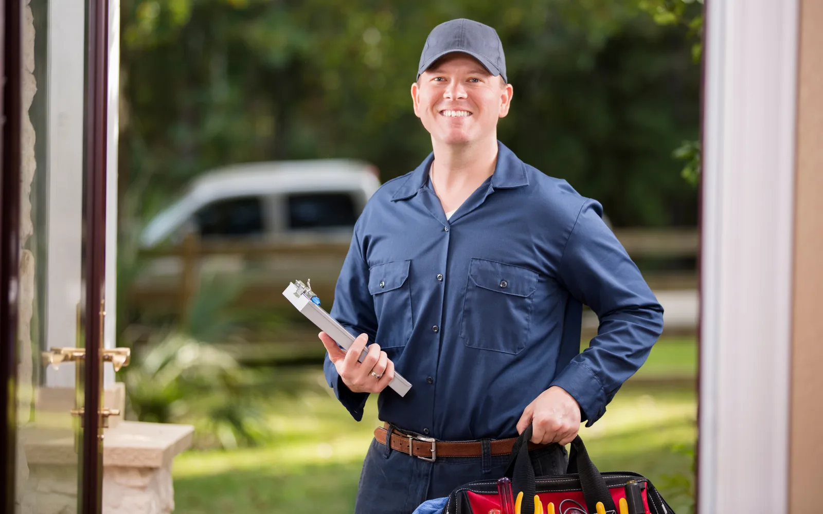 Commercial cleaning: a uniformed technician standing at a door with a clipboard in one hand and a tool bag in the other, a work van parked on the drive behind him.