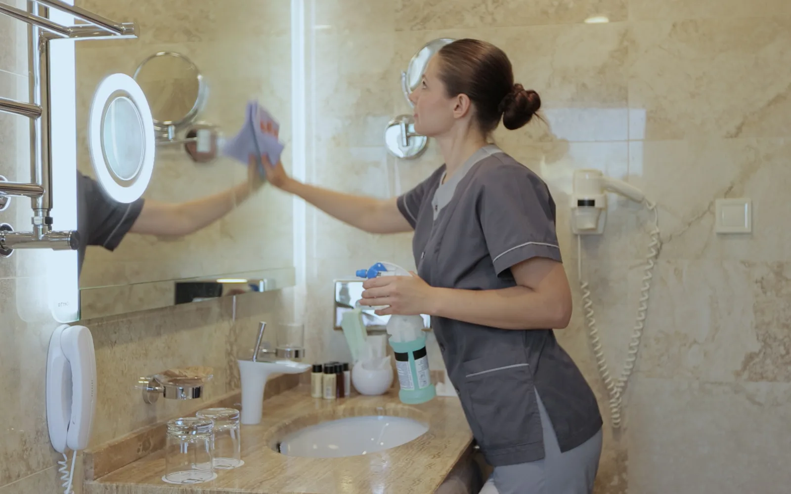 Commercial cleaning: a housekeeper in a grey tunic wiping a mirror with a lilac microfibre cloth above a marble washroom counter, glasses and amenity bottles set out below.