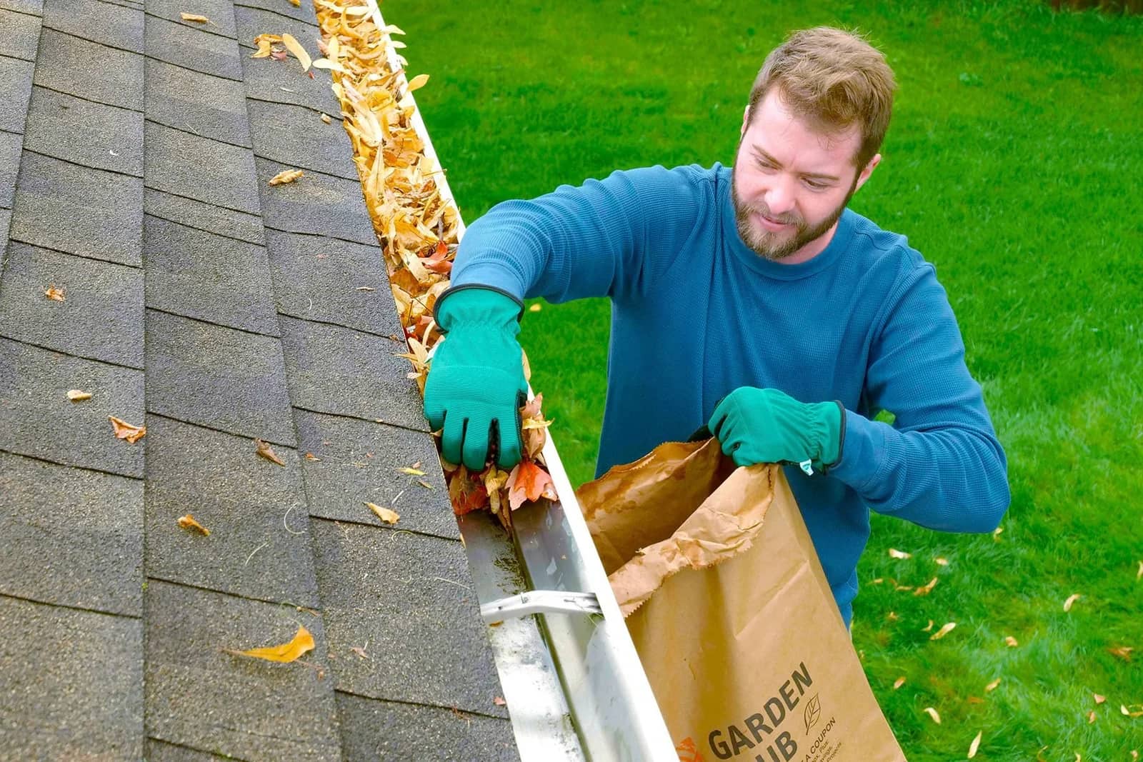 Gutter cleaning: a worker at the top of a ladder lifting leaf litter out of a roof gutter into a paper garden sack.