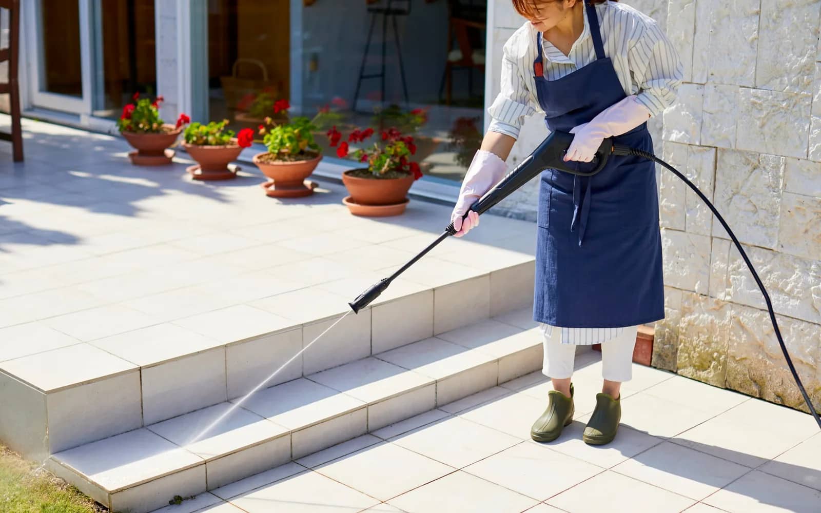 Power washing: a lance played across the treads of a pale stone stair and the paving at its foot, potted geraniums lined along the wall behind.