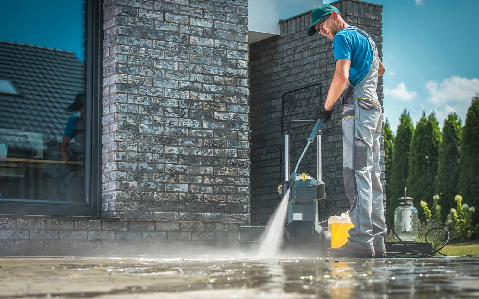 Power washing: an operator running a wheeled surface cleaner across paving beside a dark brick house, spray driving out from under the head and the slab wet behind it.