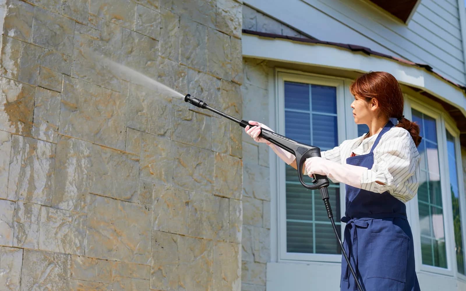 Power washing: a lance held up to a buff stone-clad wall, the spray fanning across the courses beside a white-framed window.