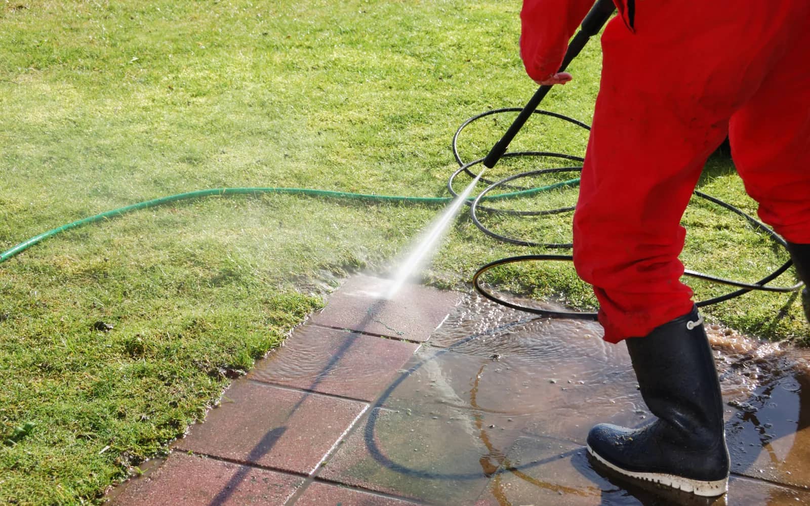 Power washing: an operator in red waterproofs washing a paved path along the edge of a lawn, water running off the slabs into the grass.