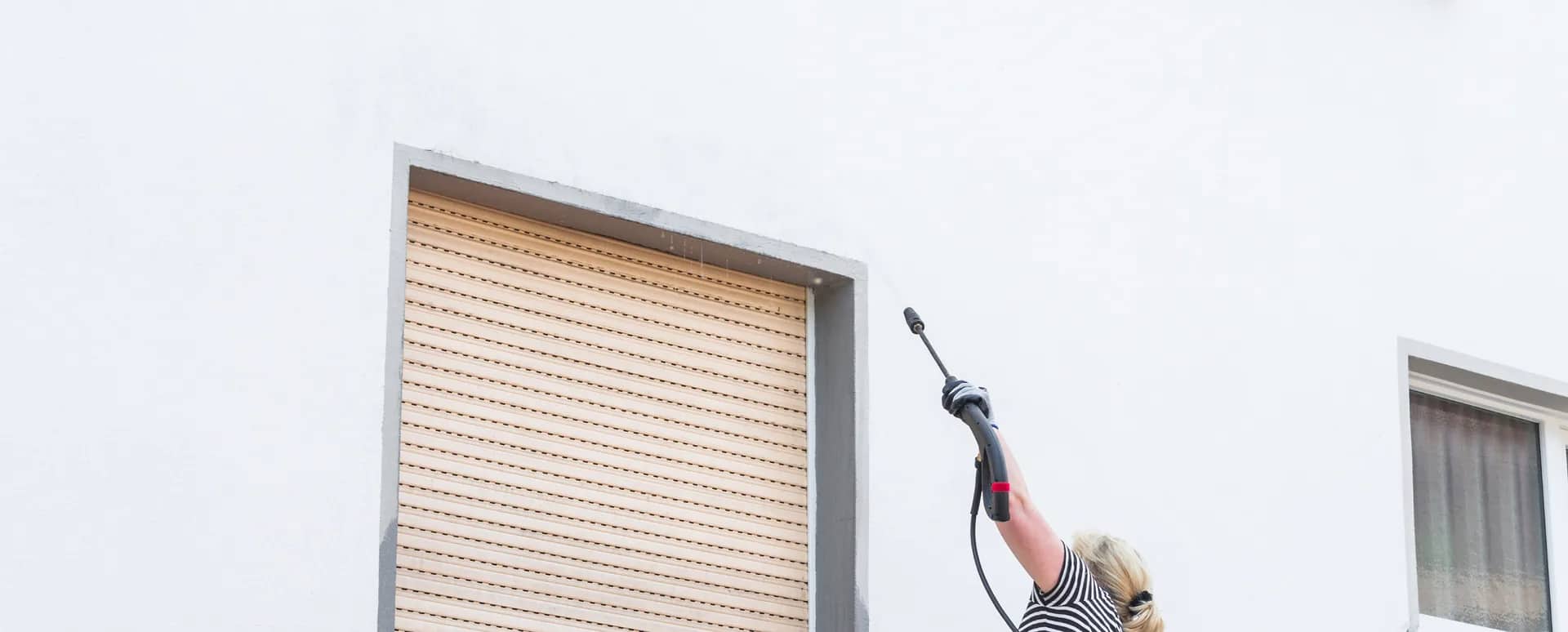 Power washing: a lance raised to the render of a white wall beside a shuttered window, the raised arm and the hose the only things breaking a plain elevation.