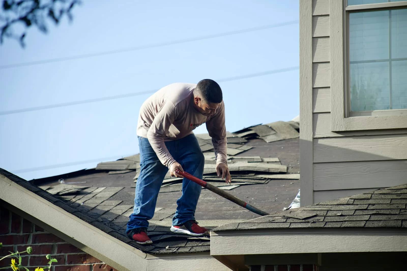 Roof cleaning: a worker standing on an asphalt shingle slope working a hand tool across the courses, the siding and window of the upper storey behind him.