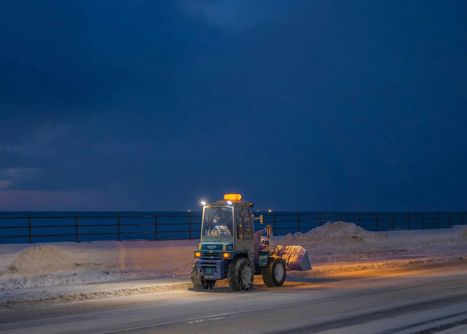 Snow removal: a loader working under its own lights at first light, a pile of cleared snow stacked at the edge of an open lot against a deep blue sky.