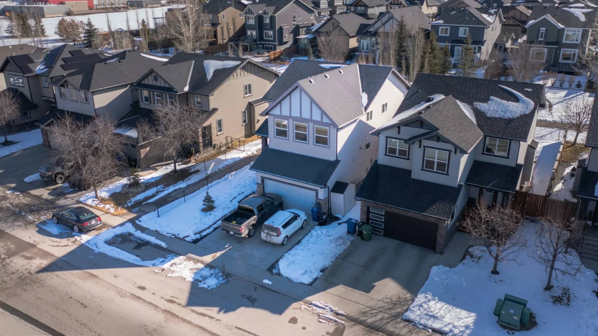 A suburban street from above after a snowfall, every driveway and path cleared to the surface while the lawns and roofs either side are still under snow.