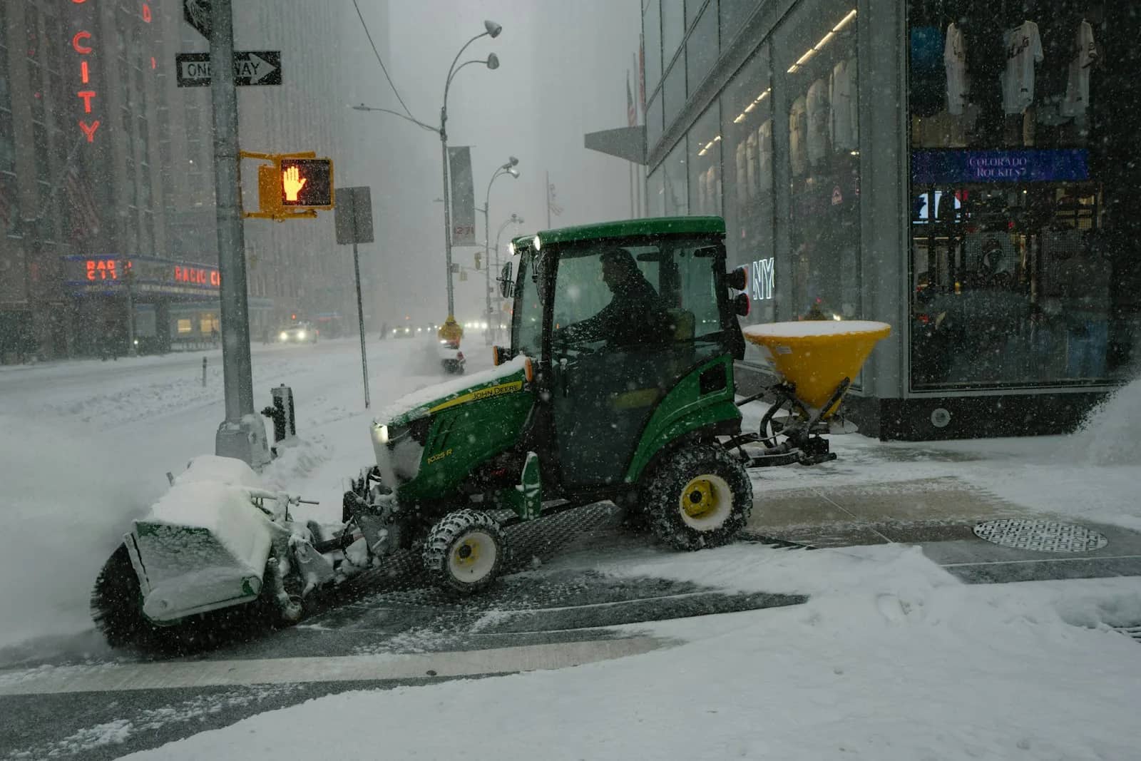 Snow removal: a compact tractor with a broom on the front and a hopper spreader on the back working a city footway through falling snow.