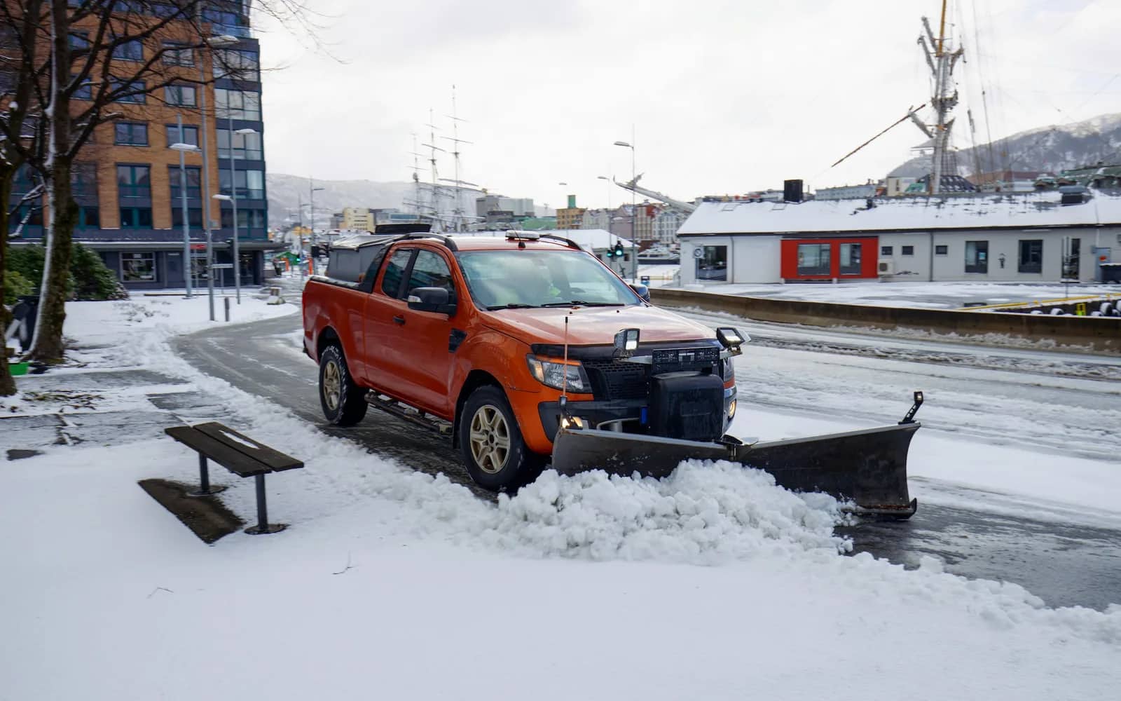 Snow removal: a pickup truck with a V-plough clearing fresh snow from a paved lane beside a bench, the cleared track wet behind it.