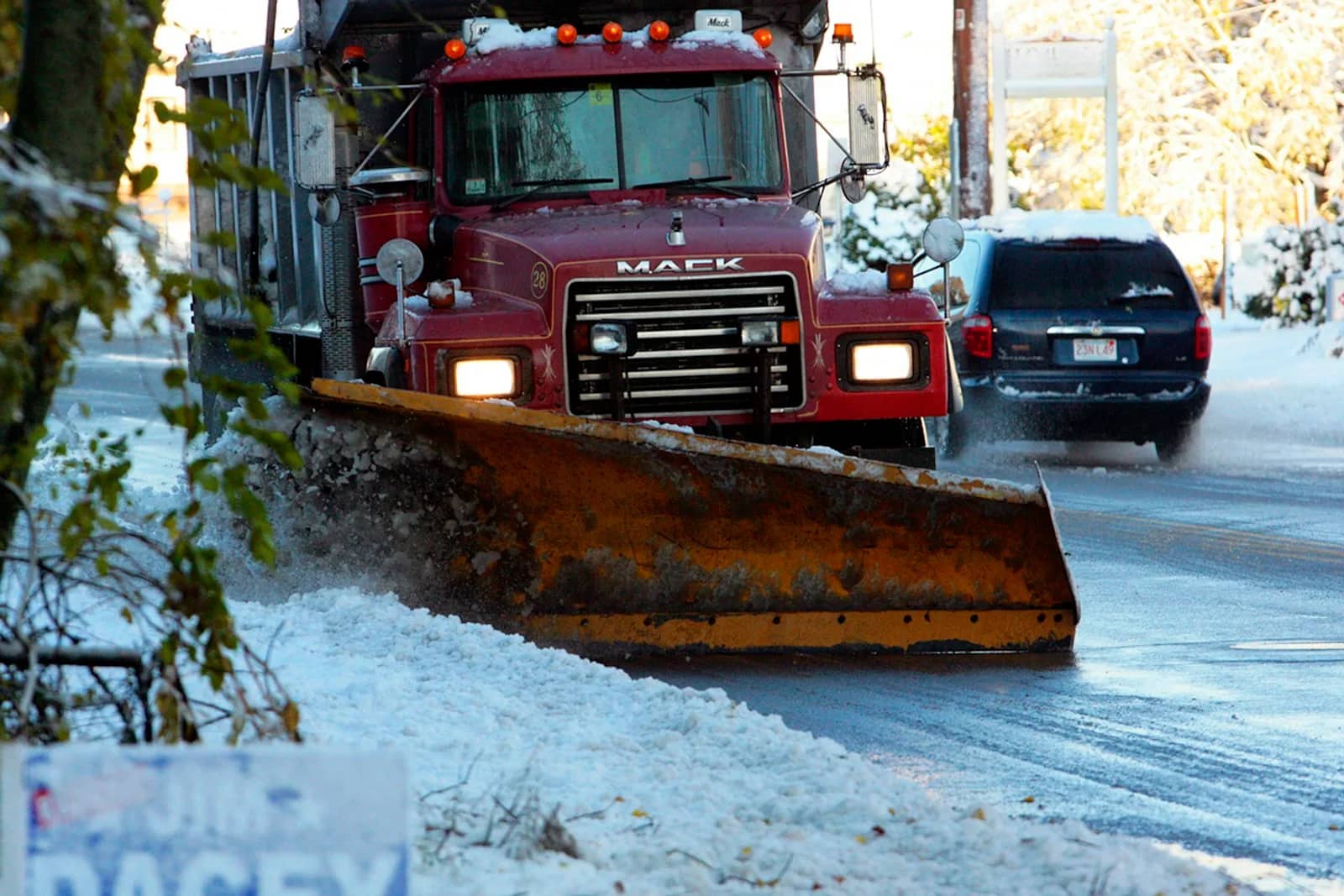 Snow removal: a plough truck with its blade angled and down, pushing a windrow of snow along a road with a car waiting behind it.