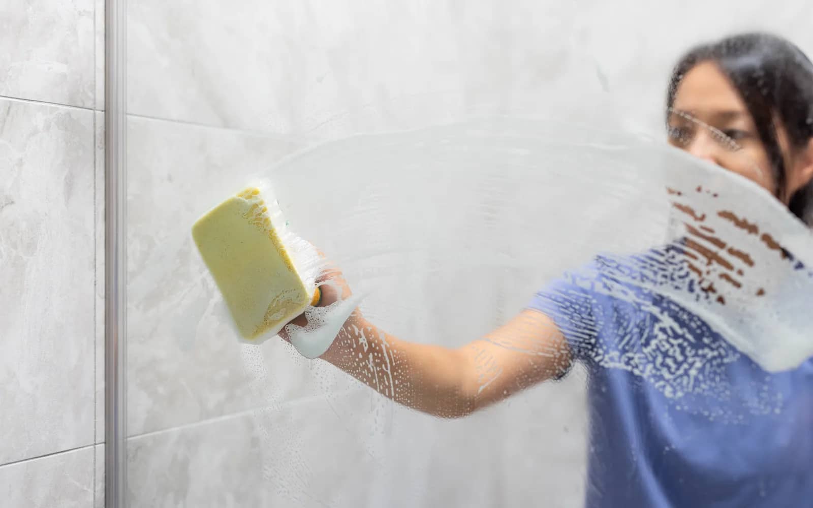 Window cleaning: a soapy sponge worked across a glass shower screen, a clear arc opening through the film with a tiled wall behind.