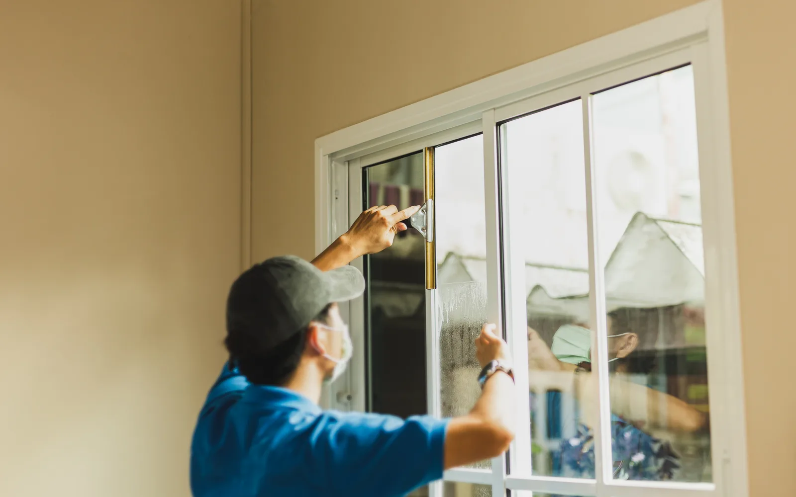 Window cleaning: a cleaner in a blue work shirt drawing a squeegee down the inside of a sliding patio door, the cleared strip sharp against the wet glass beside it.