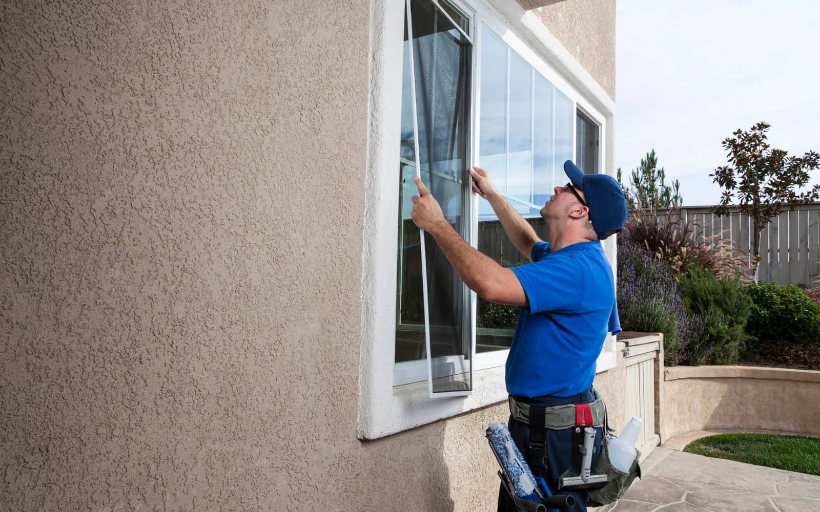 Window cleaning: a technician in a blue polo, squeegee and scrubber on the tool belt, lifting an insect screen out of a window in a stucco wall.