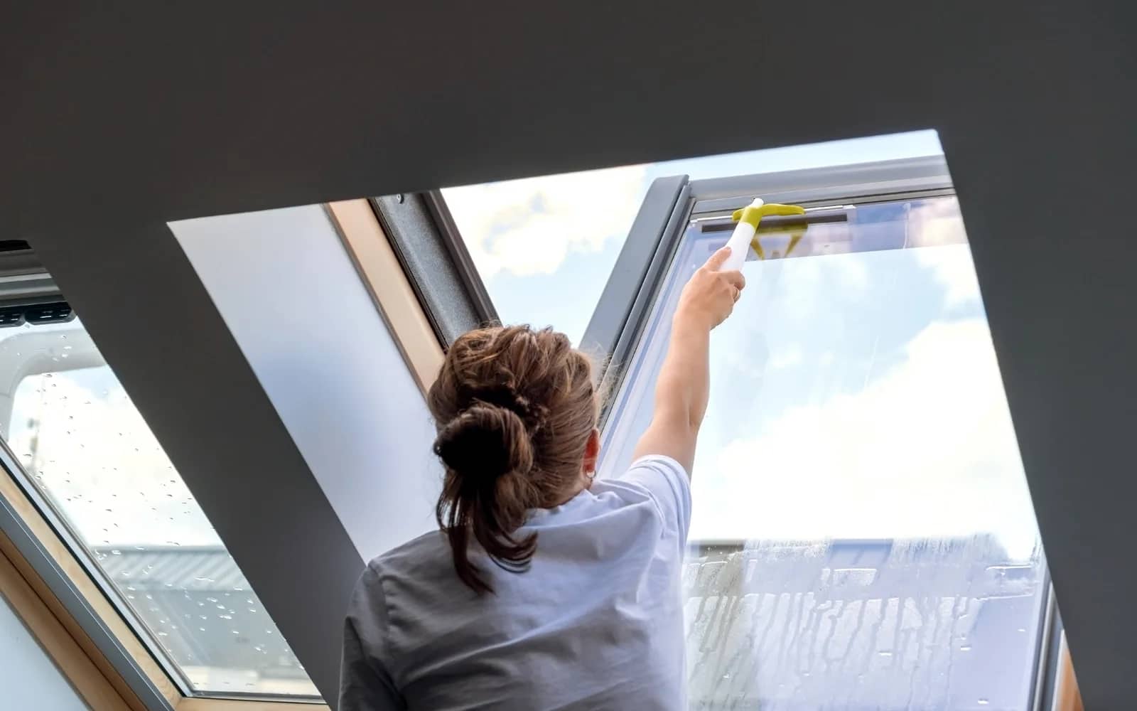 Window cleaning: a cleaner reaching up to wash a pitched-roof skylight from inside the room, a spray-and-squeegee tool drawn down the wet glass.