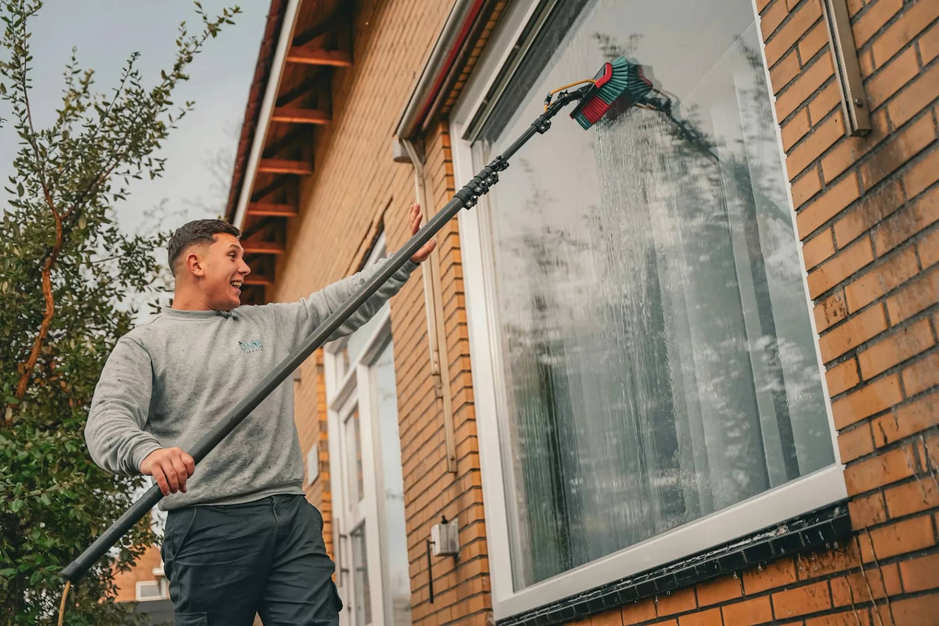 Soft washing: an operative running a water-fed pole with a soft brush head down the exterior of a brick house, water sheeting off the surface.