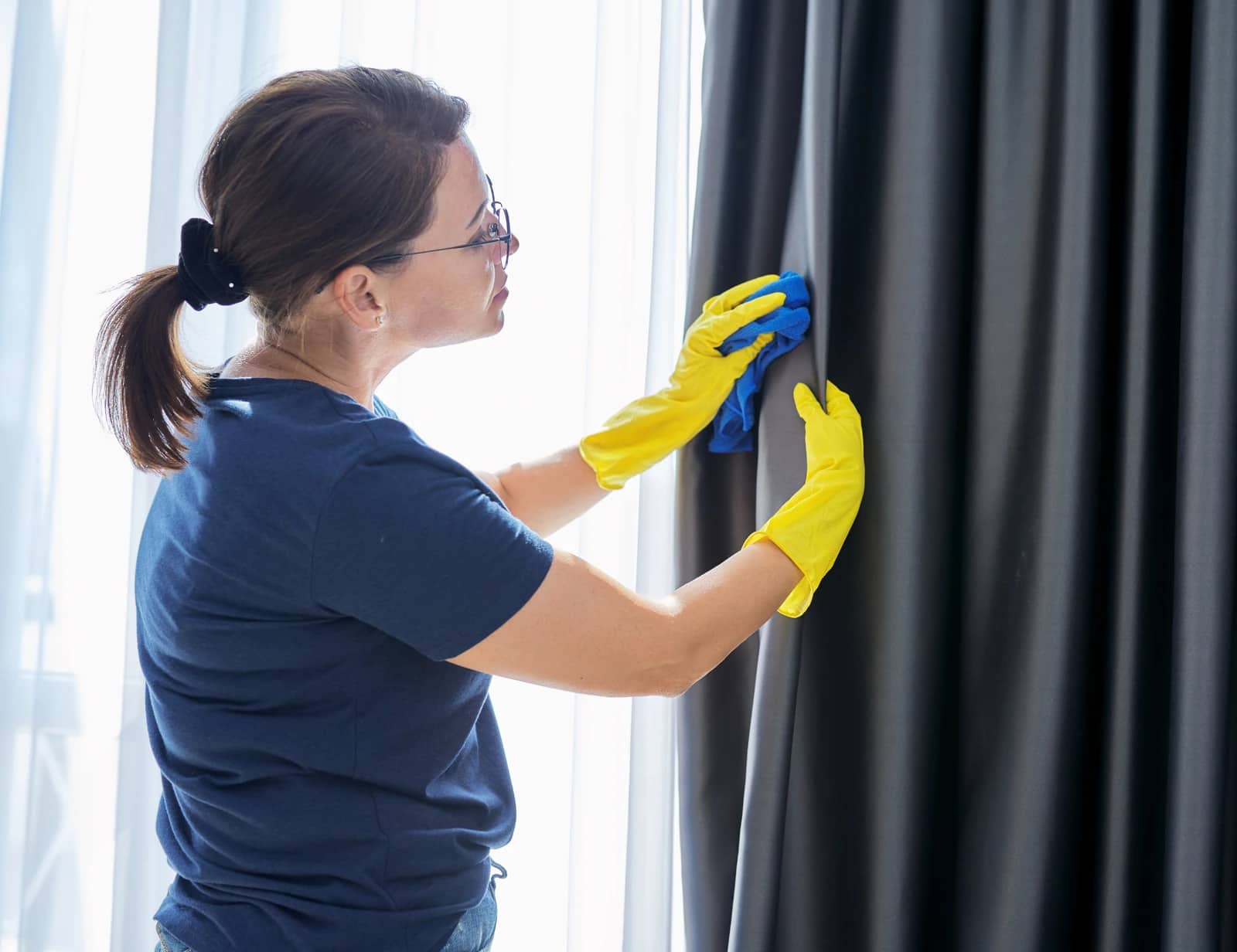 A cleaner in rubber gloves carefully wiping down a blackout curtain beside a sunlit window.