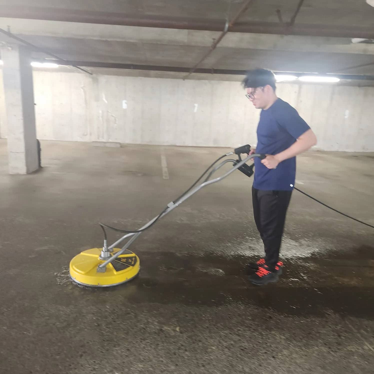 A RainCity technician guiding a yellow surface cleaner across the concrete floor of an underground parkade, the freshly washed strip dark against the dry slab.
