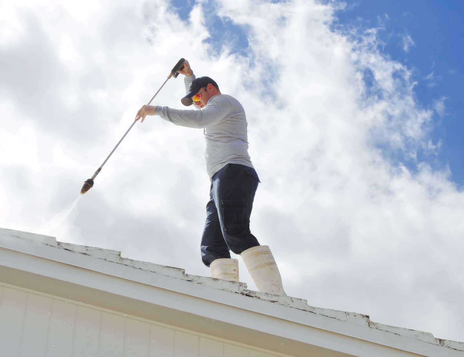 A RainCity technician silhouetted against a bright sky, running a pressure-washing wand along a rooftop edge.