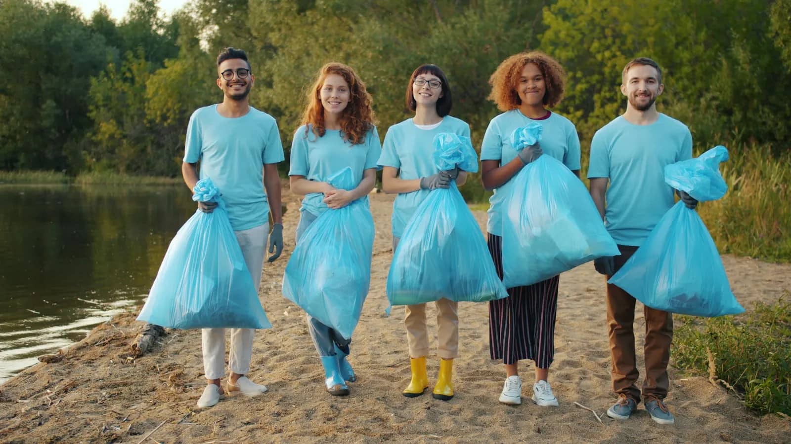 Five volunteers in matching light-blue shirts standing on a sandy path, each holding a full blue rubbish bag.