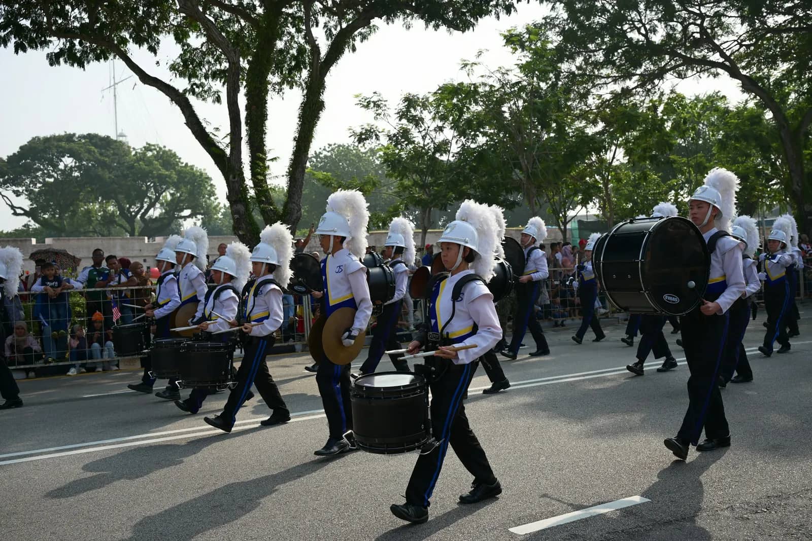 A drumline in white uniforms and plumed hats marching down a tree-lined street past spectators.