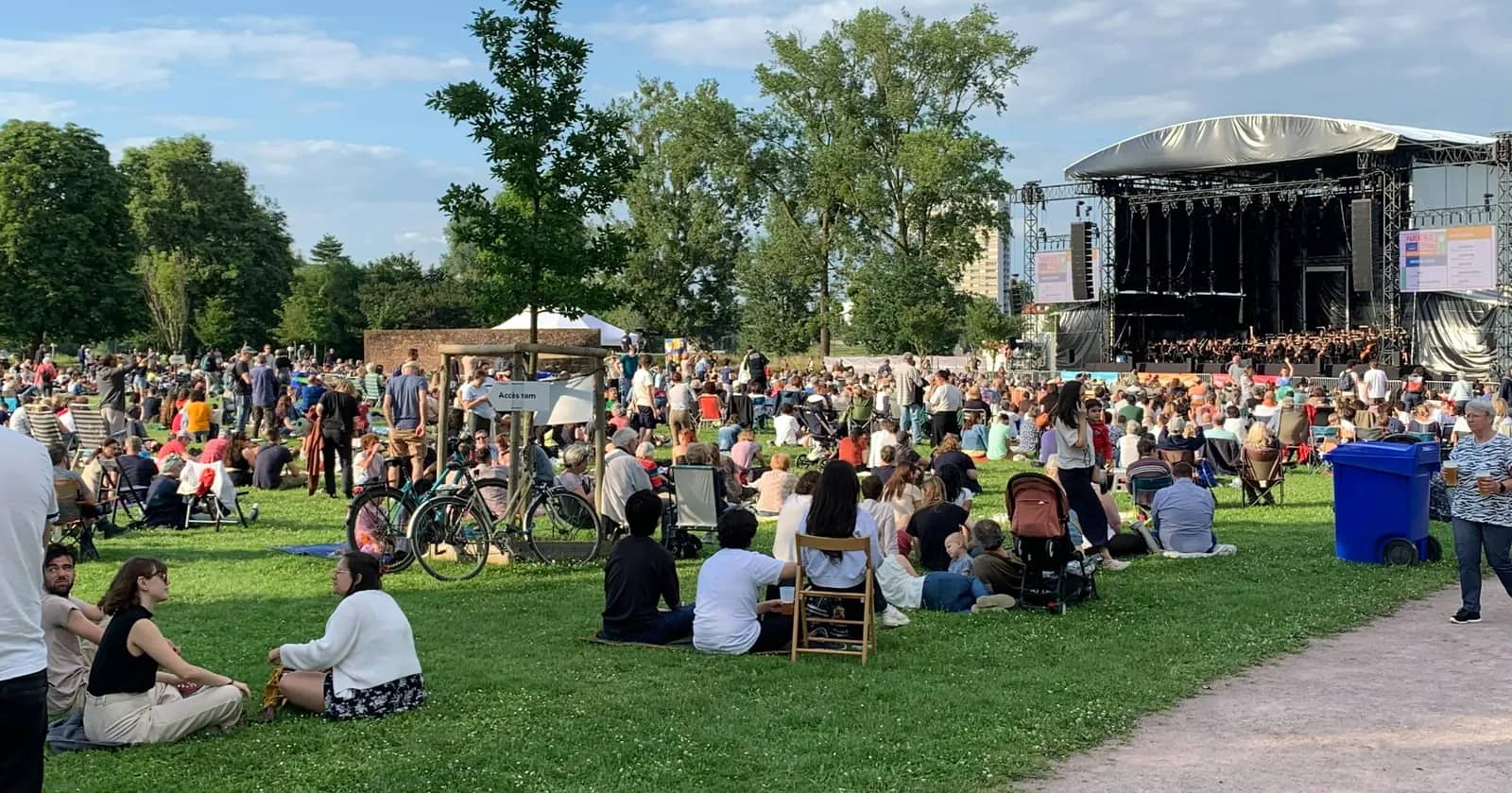 A crowd sitting and standing on the grass of a tree-lined park in front of an outdoor stage on a summer afternoon.