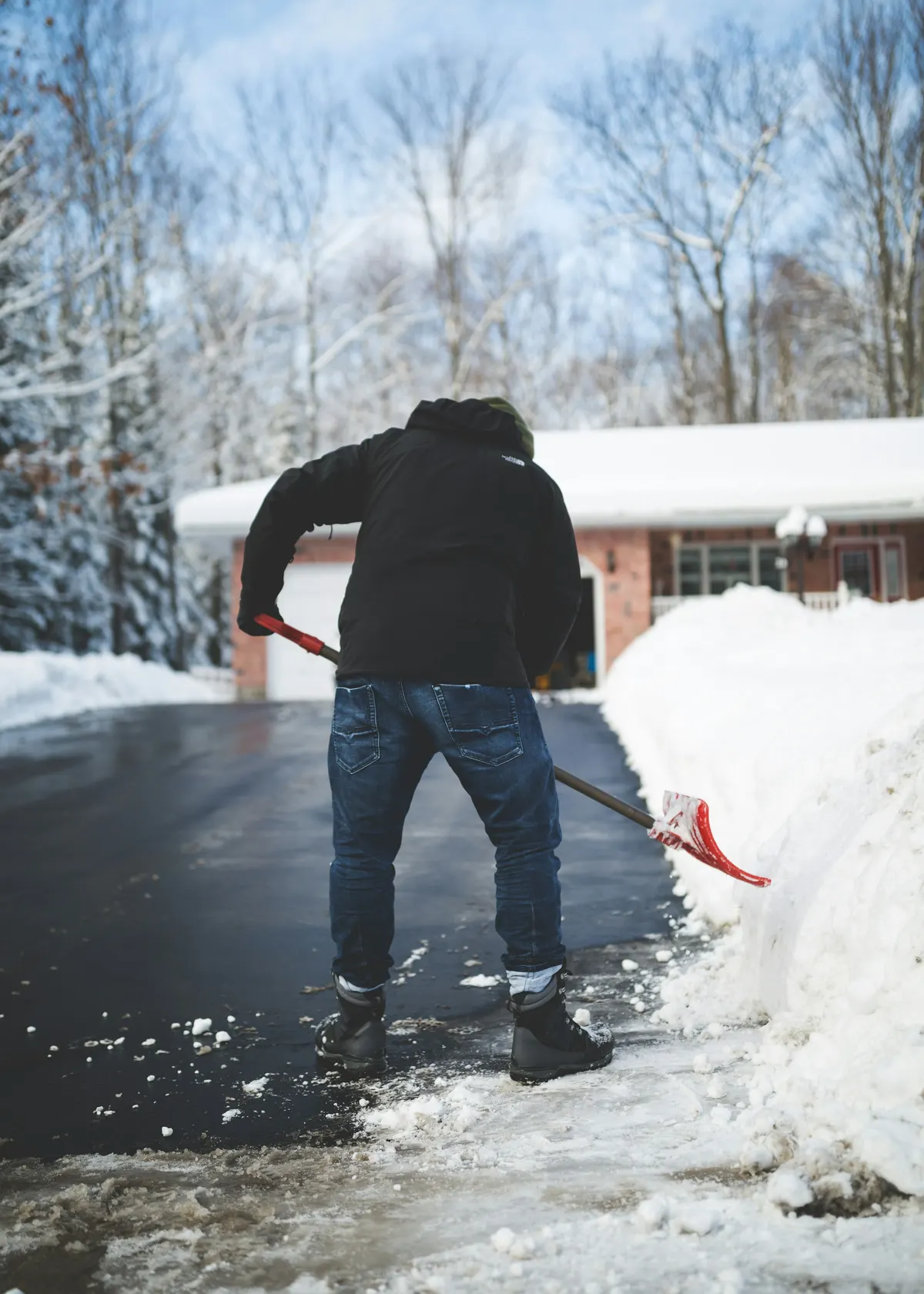 A person in a dark jacket shovelling a deep snowfall off a driveway in front of a house.