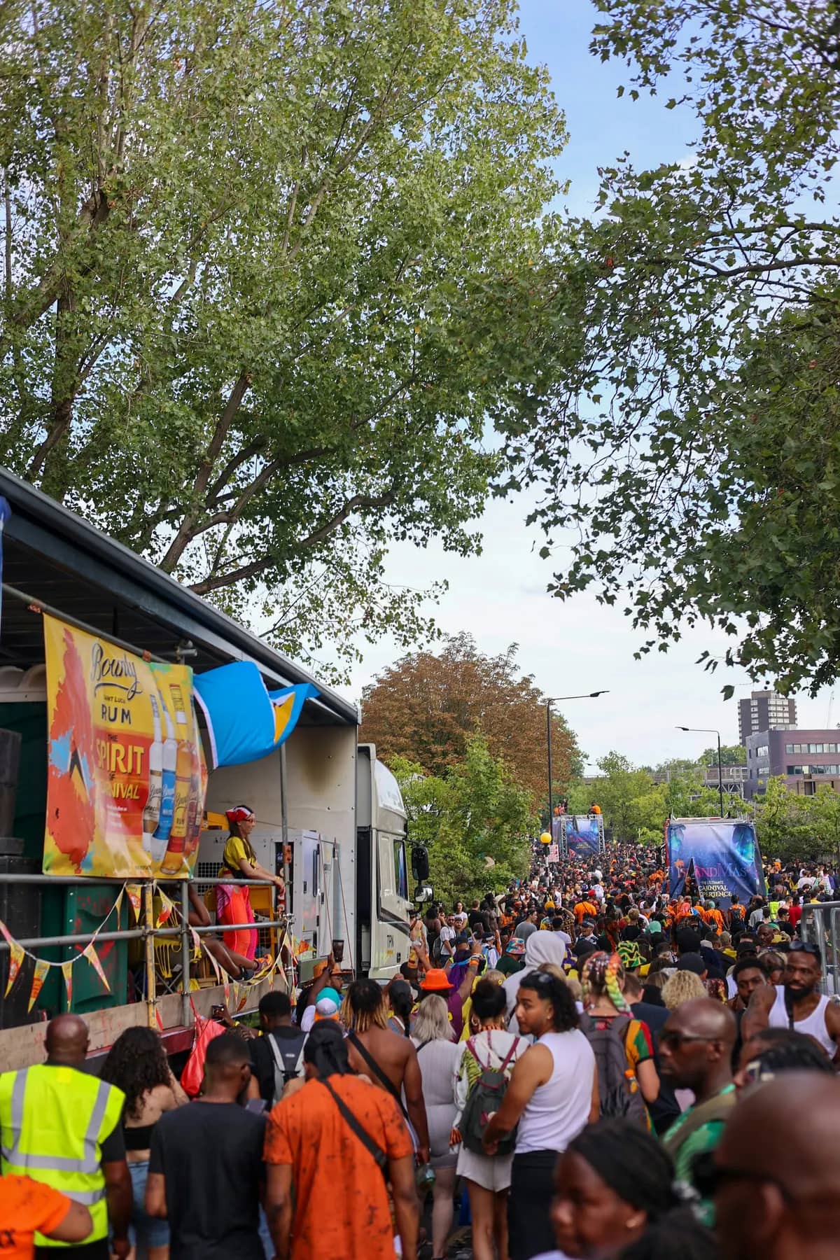 A street festival under autumn trees, with a vendor tent and bright banners in the foreground and a crowd filling the road beyond.