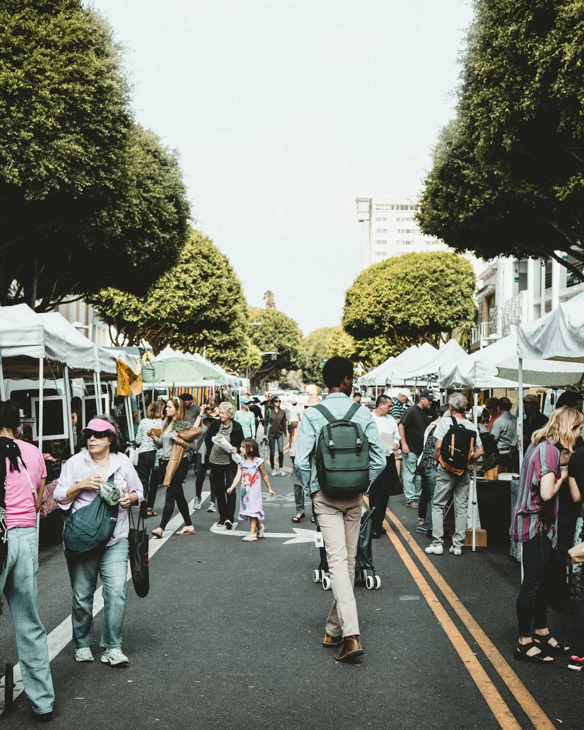 Shoppers walking between two rows of white market tents on a tree-lined street on a bright day.