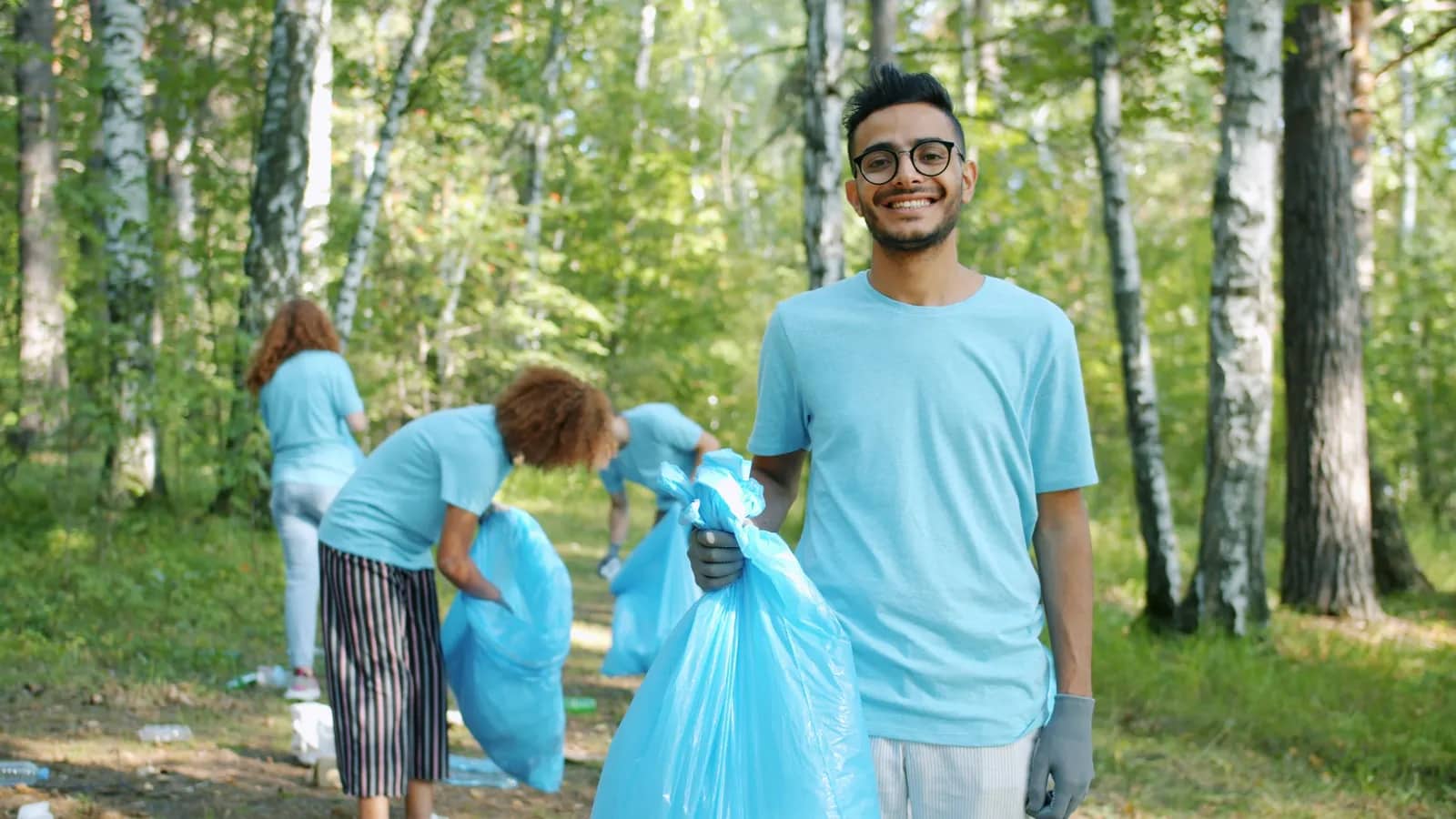 Volunteers in light-blue shirts filling blue rubbish bags along a wooded path, one of them smiling towards the camera.